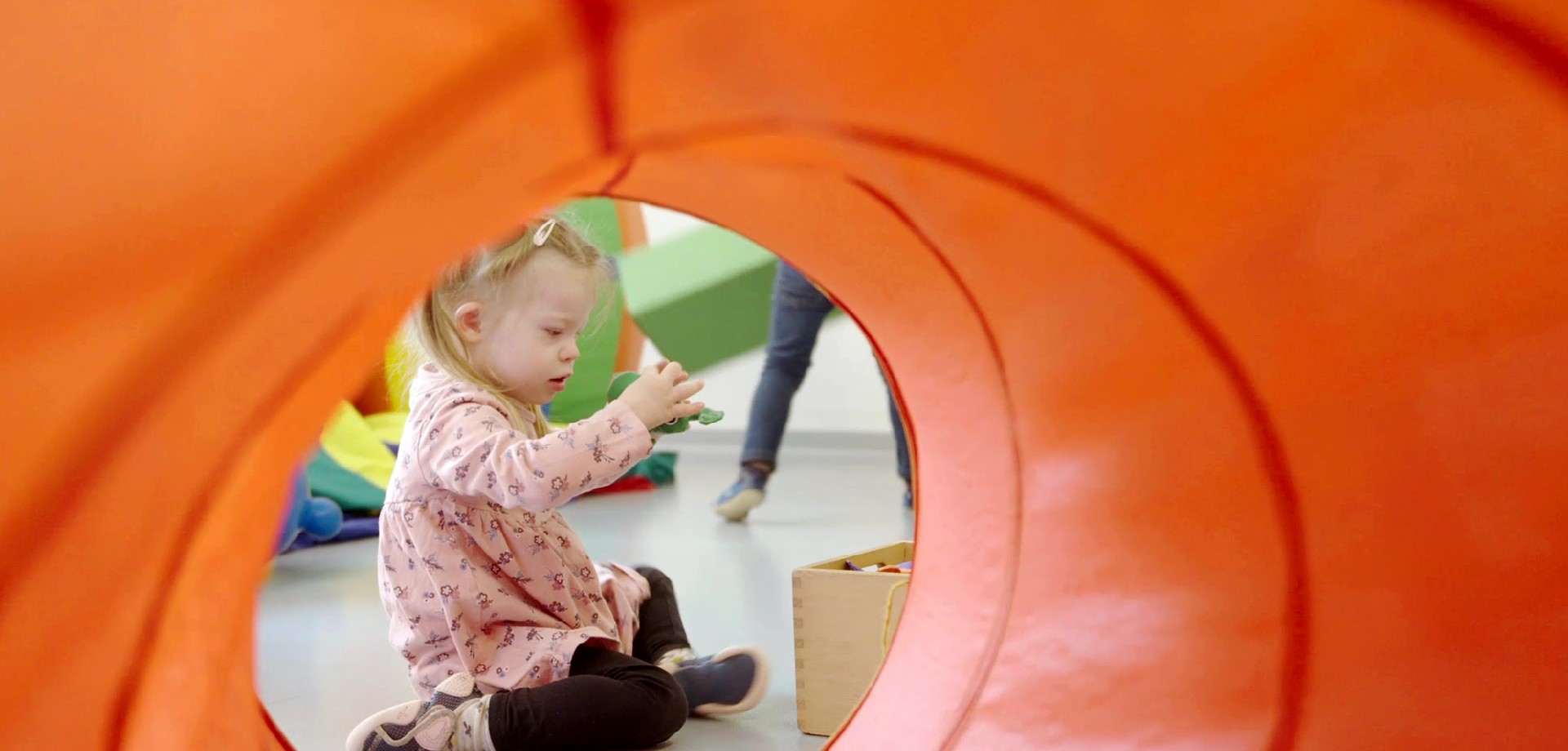 Bild: Man sieht ein kleines Mädchen mit blonden Haaren beim Spielen durch einen Krabbeltunnel.