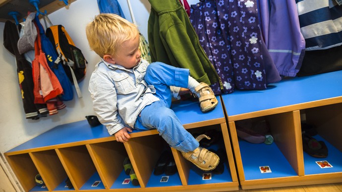 Kind sitzt auf einer Bank an der Garderobe eines Kindergartens und zieht sich die Schuhe an